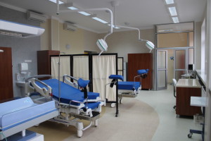 Zdjęcie bloku porodowego. Łóżka szpitalne, oddzielone parawanami. Nad nimi lampy szpitalne.