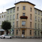 Na zdjęciu budynek starostwa powiatowego w rynku w Dzierżoniowie. Widać budynek w kolorze żółto-zielonym. Przed budynkiem zaparkowane samochody.