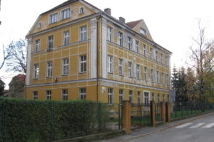 Budynek po szkole w Pieszycach. Obiekt w kolorze żółtym z wieloma oknami, otoczony płotem. Przed budynkiem ulica i przejście dla pieszych.