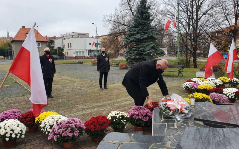Na zdjęciu moment składania kwiatów pod pomnikiem przez starostę. U stóp pomnika kwiaty, a po obu stronach flagi. W tle dwie osoby i budynki mieszkalne.