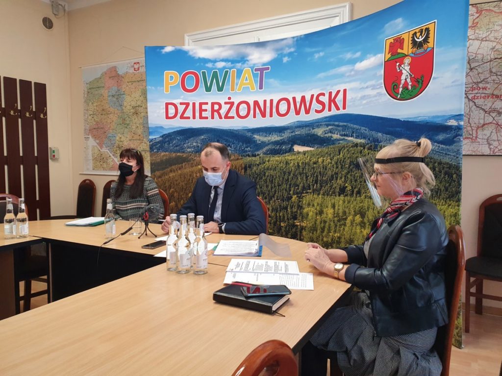Na zdjęciu trzy osoby siedzące w maseczkach ochronnych za stołem konferencyjnym. W tle ścianka reklamowa powiatu dzierżoniowskiego.