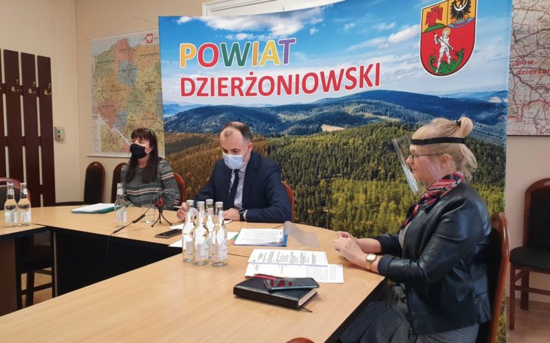 Na zdjęciu trzy osoby siedzące w maseczkach ochronnych za stołem konferencyjnym. W tle ścianka reklamowa powiatu dzierżoniowskiego.