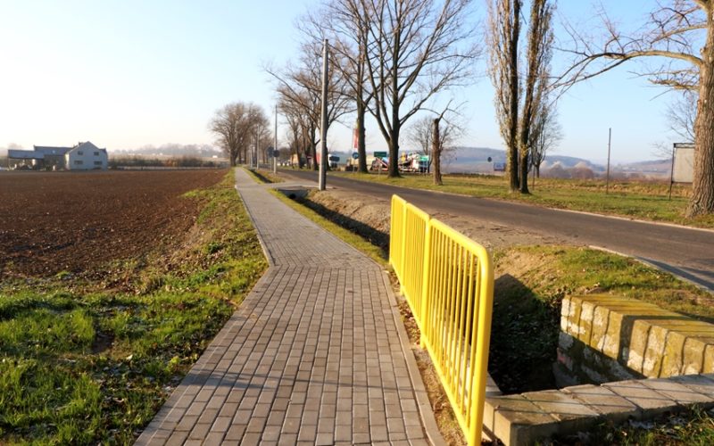 Na głównym planie chodnik z żółtą barierką ochronną. Po prawej stronie droga, a po lewej pole uprawne.
