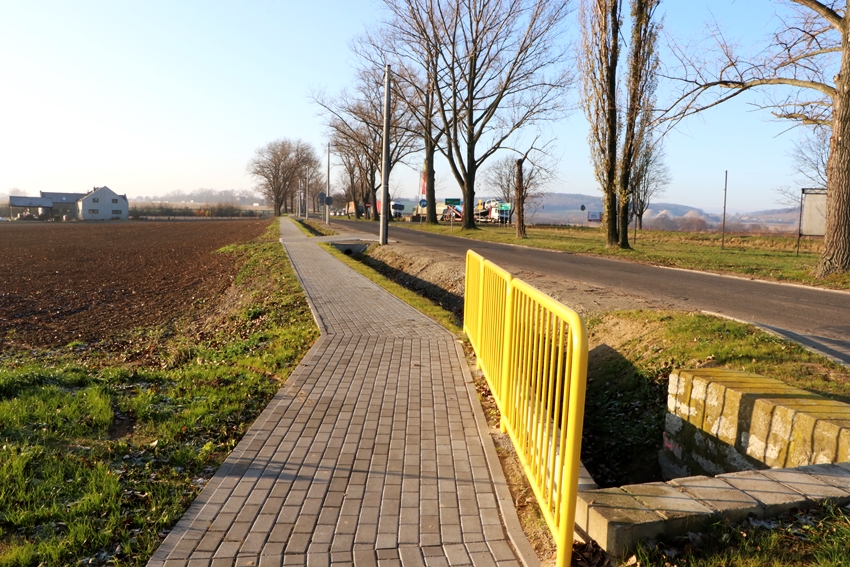 Na głównym planie chodnik z żółtą barierką ochronną. Po prawej stronie droga, a po lewej pole uprawne.