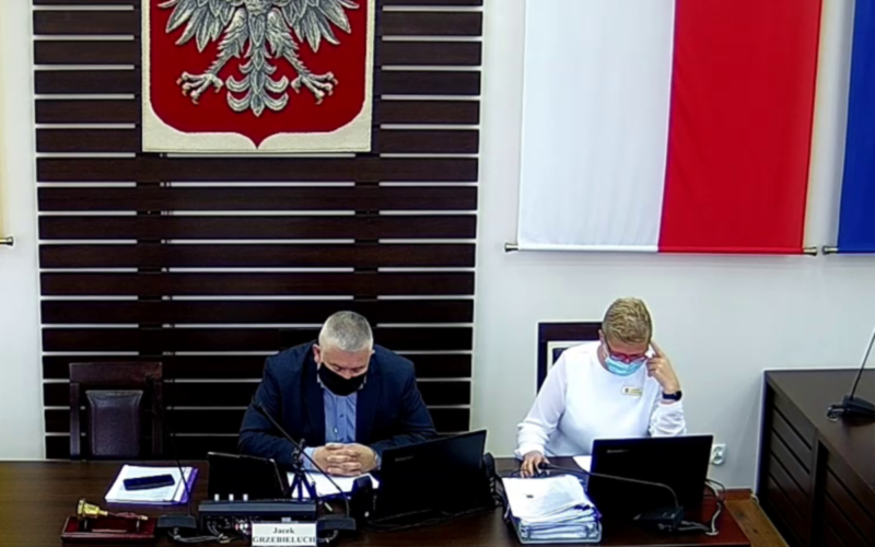 Zdjęcie sali sesyjnej Rady Powiatu Dzierżoniowskiego. Na zbliżeniu widać dwie osoby siedzące za stołem prezydialnym. Widzimy przewodniczącego Rady Powiatu oraz pracownika Starostwa. W tle widać flagę i godło RP.