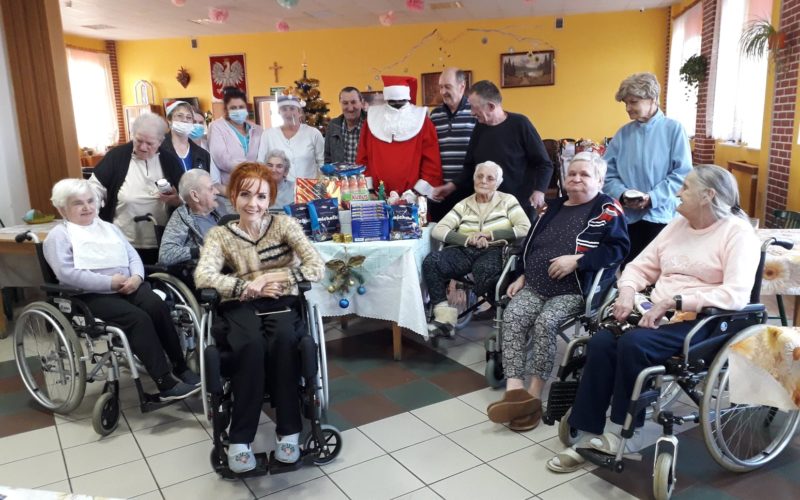 Zdjęcie grupowe mieszkańców DPS z Mikołajem. Na pierwszym planie osoby siedzą na wózkach inwalidzkich w pobliżu stolika z prezentami. Na drugim planie stoją podopieczni i pracownicy DPS z Mikołajem