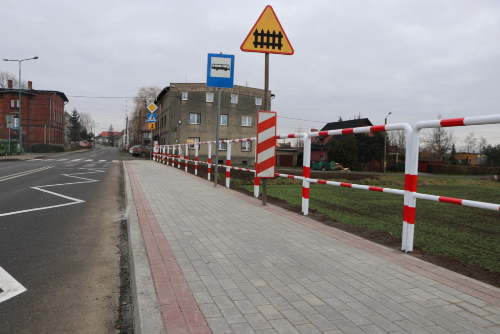 Na zdjęciu nowy chodnik z kostki betonowej wraz z barierkami ochronnymi i znakami drogowymi. W tle widać budynek wielorodzinny