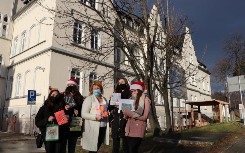 Na pierwszym planie 4 uczennice i nauczycielka trzymają kartki świąteczne. W dalszym planie budynek szpitala powiatowego w dzierżoniowie