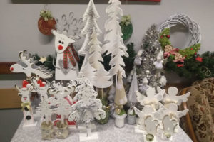 Zdjęcie przedstawia ozdoby świąteczne stojące w grupie na stoliku. Widać ręcznie wykonane z drewna ozdoby w kolorze białym, przedstawiające choinki, renifery, aniołki.