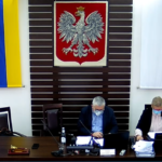 Zdjęcie sali sesyjnej Rady Powiatu Dzierżoniowskiego. Na zbliżeniu widać dwie osoby siedzące za stołem prezydialnym. Widzimy przewodniczącego Rady Powiatu oraz kierownika Biura Rady Powiatu. W tle widać flagę i godło RP.