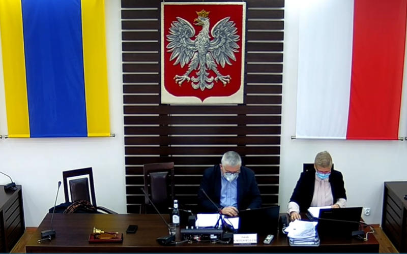 Zdjęcie sali sesyjnej Rady Powiatu Dzierżoniowskiego. Na zbliżeniu widać dwie osoby siedzące za stołem prezydialnym. Widzimy przewodniczącego Rady Powiatu oraz kierownika Biura Rady Powiatu. W tle widać flagę i godło RP.