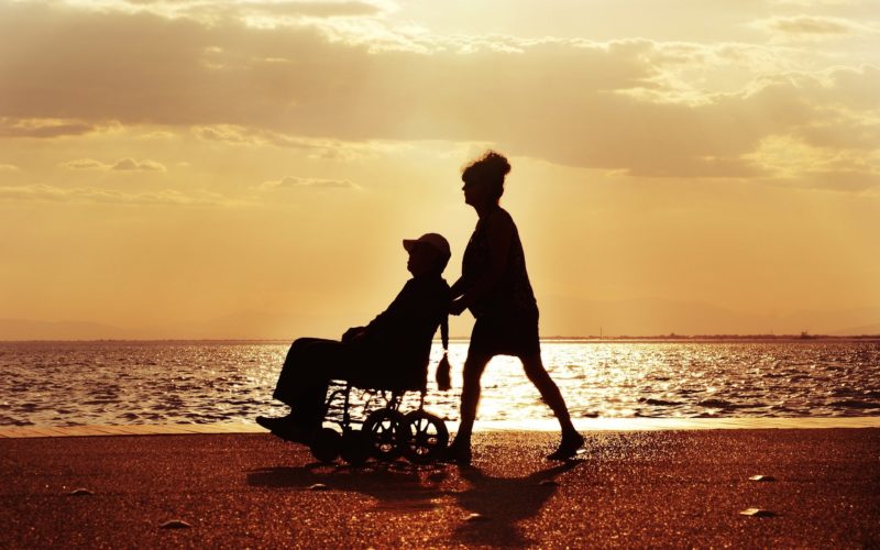 Zdjęcie plaży w czasie zachodzącego słońca. Brzegiem spaceruje osoba, która pcha wóżek z osobą niepełnosprawną. W tle morze