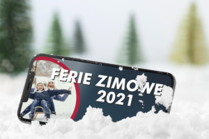 Banner informujący o Feriach Zimowych 2021 roku. Grafika przedstawiająca telefon komórkowy wrzucony w śnieg, na którego ekranie wyświetlana jest informacja o feriach, a w lewym rogu ekranu zdjęcie ilustrujące zabawę dziecka z opiekunką na sankach. W tle rozmazane świerki.