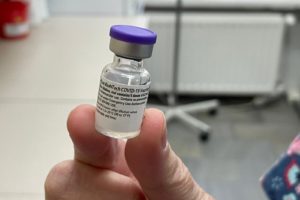 na pierwszym planie ampułka ze szczepionką przeciwko covid-19 trzymana w palcach. w tle najprawdopodobniej gabinet lekarski