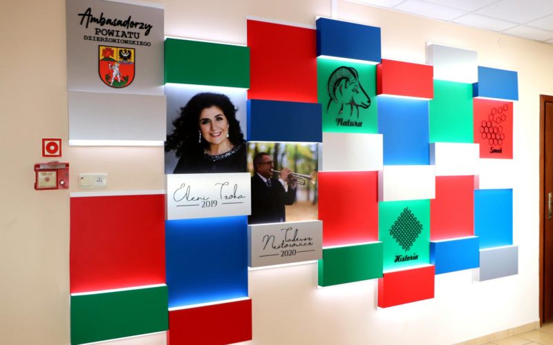 Zdjęcie ścianki promującej ambasadorów powiatu. Ściana składająca się z bloków kolorowych z fotografiami wyróżnionych osób.