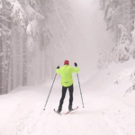 Zdjęcie przedstawiające narciarza biegowego na trasie biegowej w ośniezonym lesie, ubranego w jaskrawo zielona kurtkę, czarne spornie i czerwoną czapkę z kijkami narciarskimi w dłoniach, z uniesionymi ramionami.