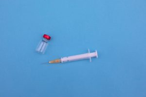 Grafika ilustrująca szczepienia. na zdjęciu widoczna biała strzykawka z igłą z nasadką koloru żółtego, oraz przeźroczysta fiolka z lekiem, z wieczkiem w kolorze czerwonym. Elementy ułożone na błękitnym tle.