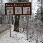 Tablica turystyczna znajdująca się na poboczu drogi w górach. Zdjęcie wykonane zimą, dookoła śnieg.
