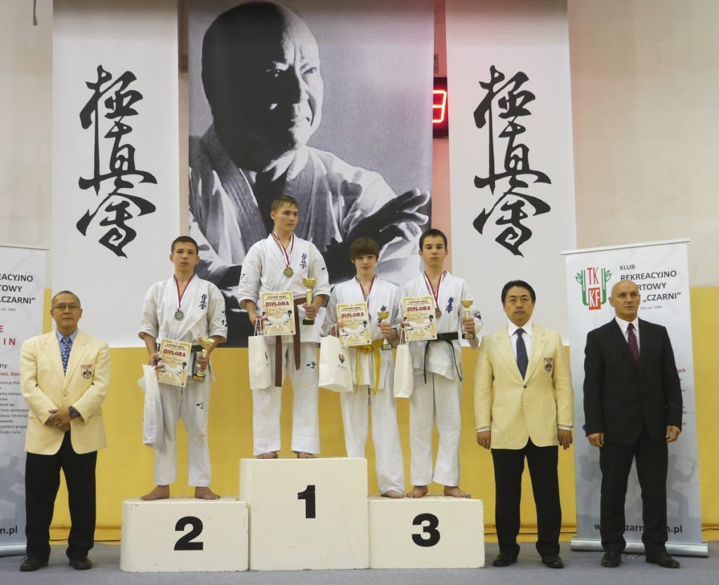 na podium stoją zawodnicy z nagrodami i w kimonach. Obok sędziowie