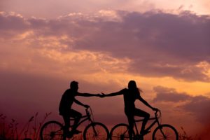 Para jadąca na rowerach i trzymająca się za ręce. W tle zachód słońca
