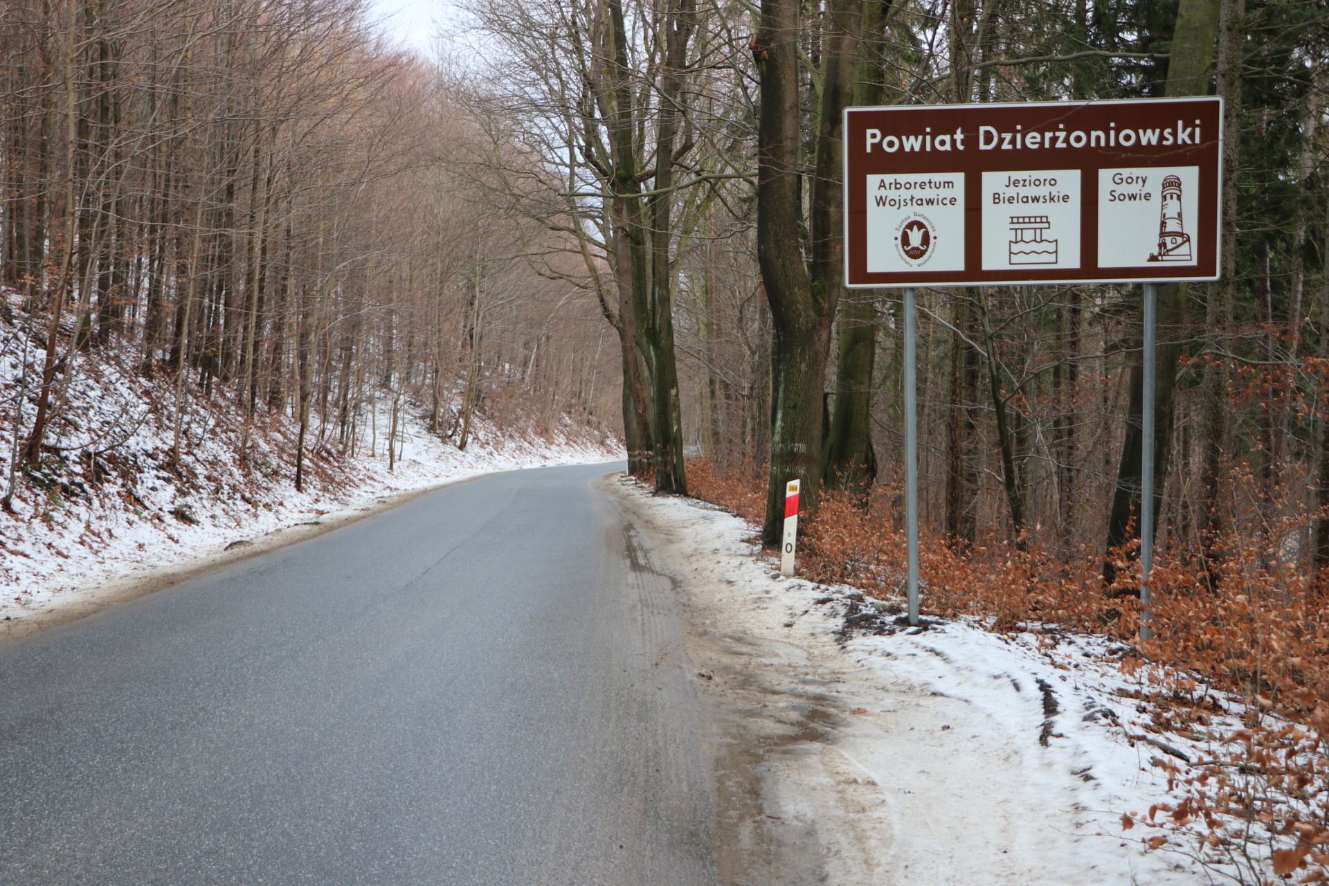 Droga dwupasmowa w górach, na poboczach śnieg, a po prawej stronie tablica informacyjna.