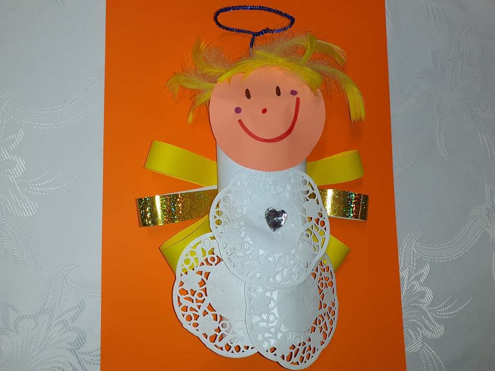Na pomarańczowej kartce aniołek w białym stroju ze skrzydłami z żółtego i złotego papieru o włosami z piór.