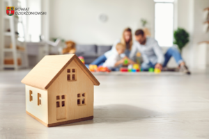 grafika przedstawiająca drewniany zabawkowy dom i rodzinę w tle