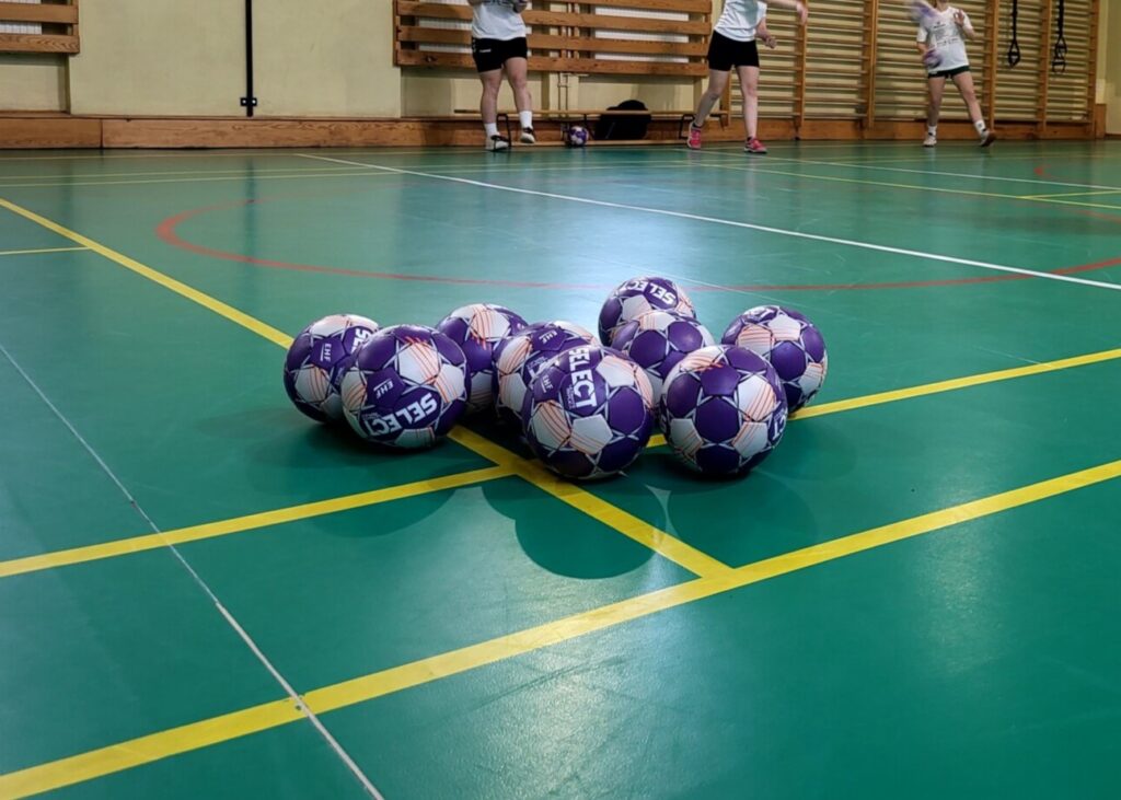 Piłki leżą na sali gimnastycznej, w tle uczniowie