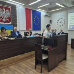 dyrektor wydziału edukacji przemawia, w tle przy stołach siedzą zarząd powiatu, przewodniczący rady i radni