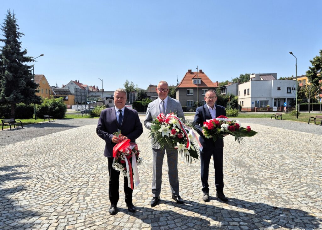 Starosta, Burmistrz Dzierżoniowa i Adam Wolniak na placu przed pomnikiem trzymają wiązanki kwiatów, w tle budynki