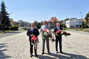 Starosta, Burmistrz Dzierżoniowa i Adam Wolniak na placu przed pomnikiem trzymają wiązanki kwiatów, w tle budynki