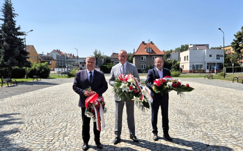 Starosta, Burmistrz Dzierżoniowa i Adam Wolniak na placu przed pomnikiem trzymają wiązanki kwiatów, w tle budynki