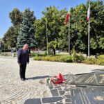Burmistrz Dzierżoniowa składa kwiaty pod pomnikiem, w tle flagi na masztach i drzewa
