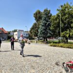 Starosta składa kwiaty pod pomnikiem, za nim stoją Burmistrz Dzierżoniowa i Adam Wolniak, w tle flagi na masztach, drzewa i budynki