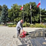 Starosta składa kwiaty pod pomnikiem, w tle flagi na masztach i drzewa