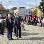 ludzie stoją z kwiatami i czekają na złożenie ich pod pomnikiem