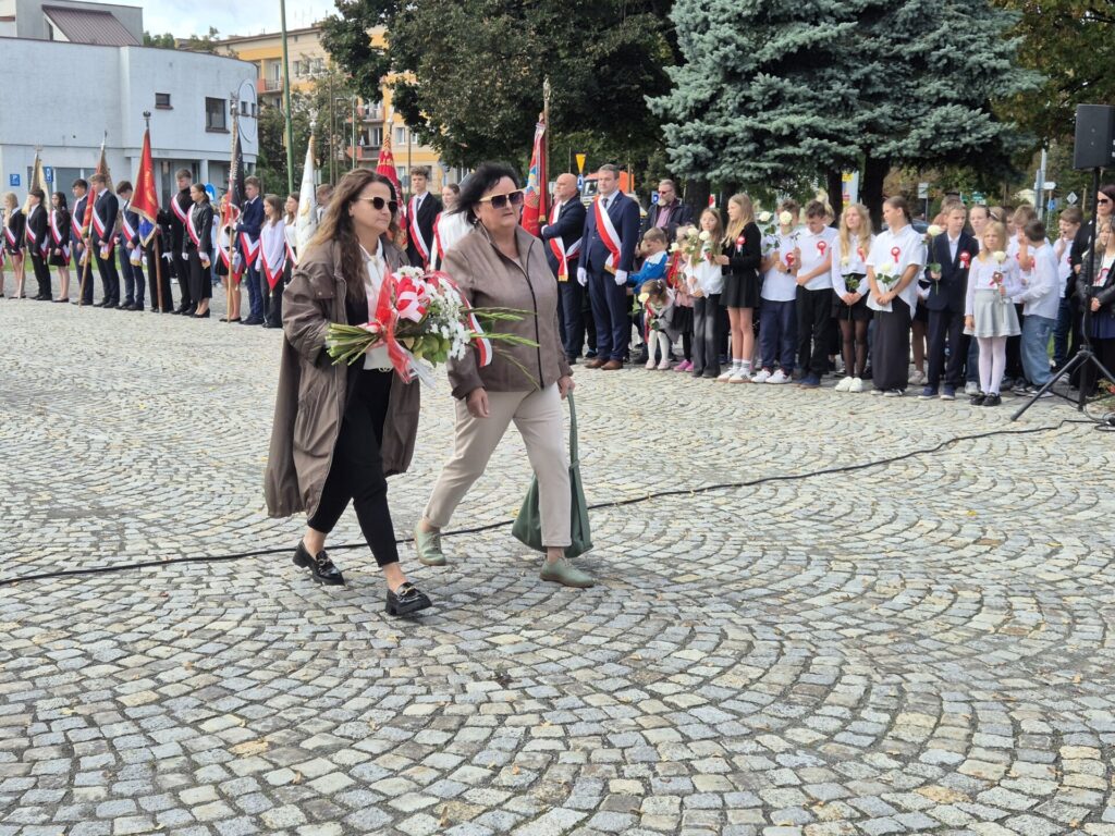 radne powiatowe idą z kwiatami, w tle poczty sztandarowe i uczniowie