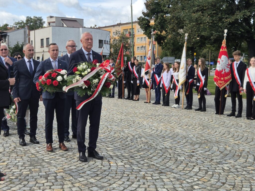 poseł czeka z kwiatami przed pomnikiem, w tle starosta i wicestarosta