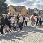 ludzie stoją z kwiatami i czekają na złożenie pod pomnikiem