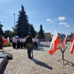 uroczyste obchody, flagi Polski stoją przy pomniku, w tle grupa ludzi