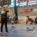 zapaśniczki podczas rywalizacji na macie, na pierwszym planie sędzia