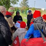 grupa młodzieży w czerwonych czapkach, stoją tyłem, obok nich troje dorosłych, w tym Burmistrz Piławy Górnej