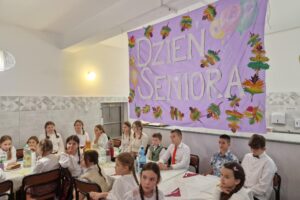 dzieci siedzą przy stoliku, w tle napis dzień seniora na różowym tle