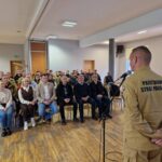 samorządowcy i strażacy siedzą na krzesłach i słuchają komendanta straży pożarnej