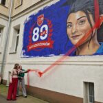 odsłonięcie czerwonej wstęgi z muralu, absolwentka w berecie na niebieskim tle, obok napis 80-lecie I LO