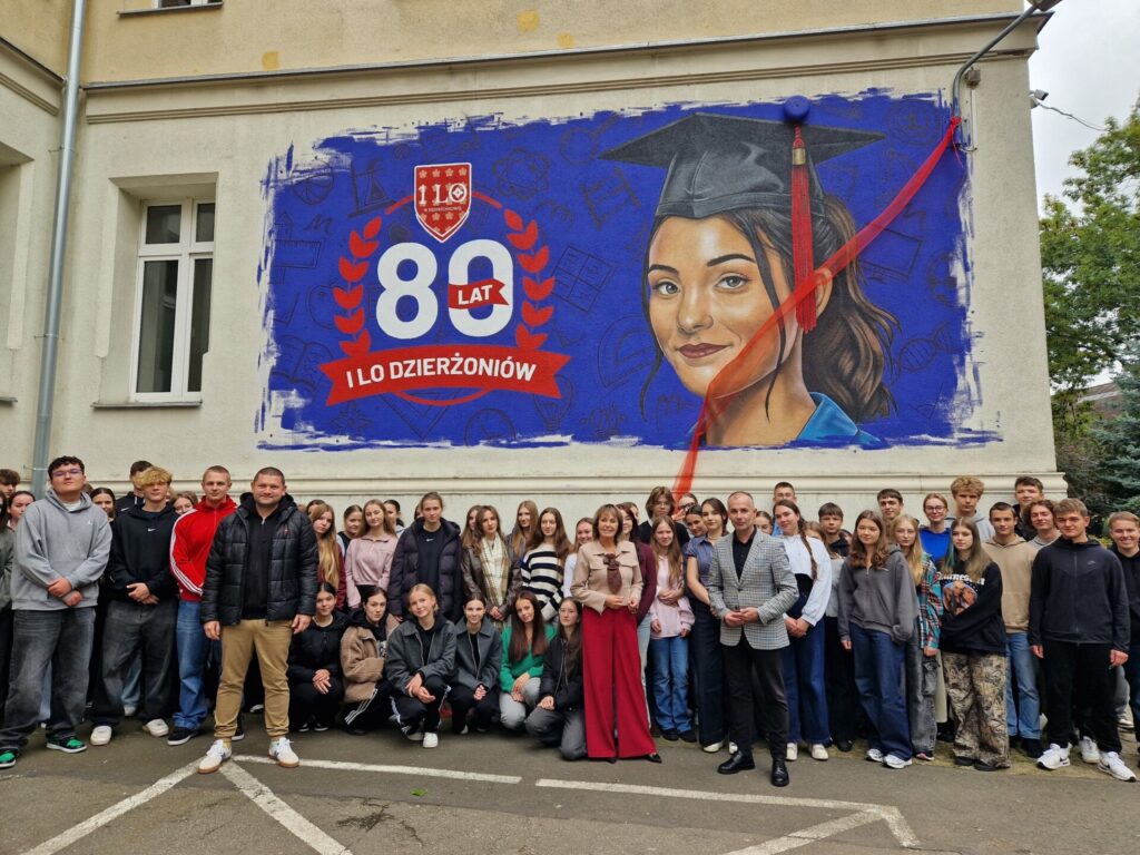 grupa uczniów wraz z dyrektor i wicestarostą stoją pod muralem z okazji 80-lecia I LO