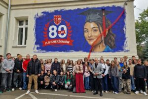 grupa uczniów wraz z dyrektor i wicestarostą stoją pod muralem z okazji 80-lecia I LO