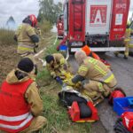 strażacy pomagają poszkodowanemu, w tle wóz strażacki