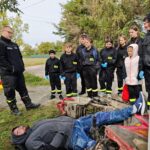 grupa młodych strażaków stoi przy poszkodowanym przygniecionym przez motor