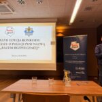 widok na ekran z nazwą konkursu, na stole szklanki, w tle puchar, policjant przemawia